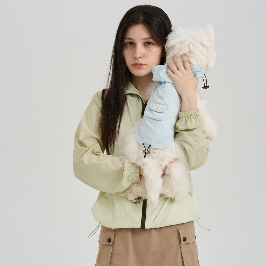 Pet parent wearing a pale green sun protection jacket while holding a white small dog in a light blue dog sun protection jacket, featuring lightweight dog apparel and coordinated outdoor style.