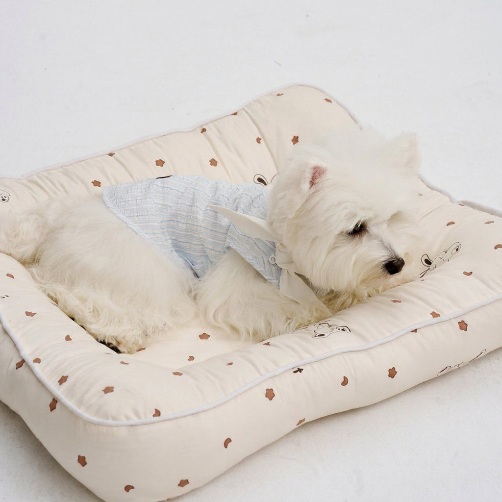 White small dog resting on a cream pet bed wearing a light blue striped dog shirt with a white tie collar, featuring comfortable dog apparel and cozy pet clothing for indoor lounging.