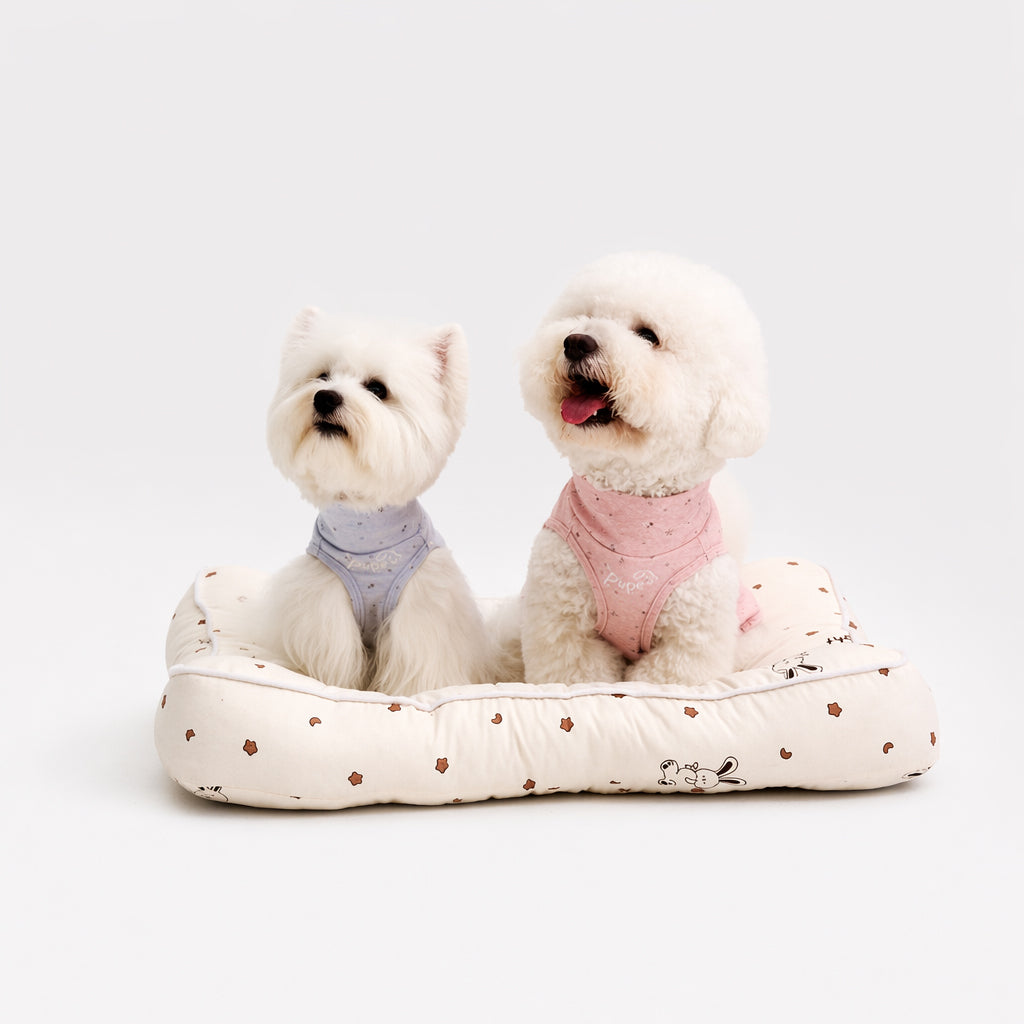 Two white small dogs sitting on a cream pet bed wearing light blue and pink speckled dog tees, featuring soft dog apparel and comfortable small dog clothes for everyday pet clothing style.