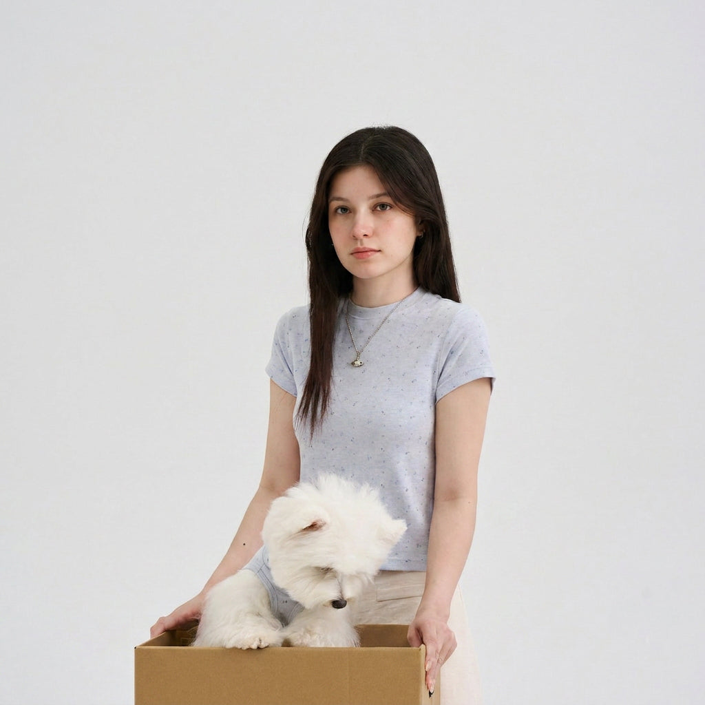 Woman wearing a light blue speckled tee standing with a white small dog in a cardboard box, featuring a matching dog shirt, soft dog apparel, and casual pet parent matching clothes.