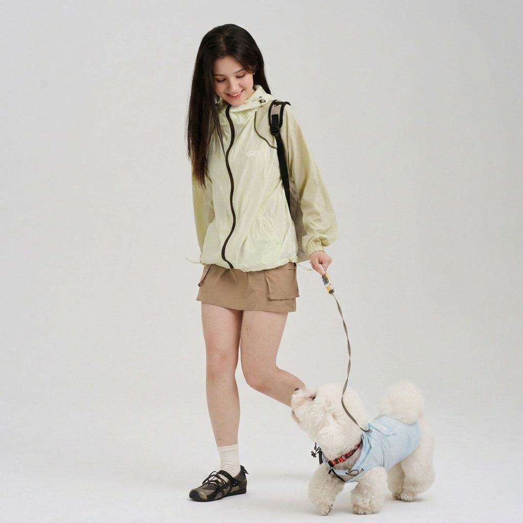 Pet parent in a pale green sun protection jacket walking a white small dog in a light blue dog sun jacket, featuring lightweight dog apparel for casual pet parent matching walks.
