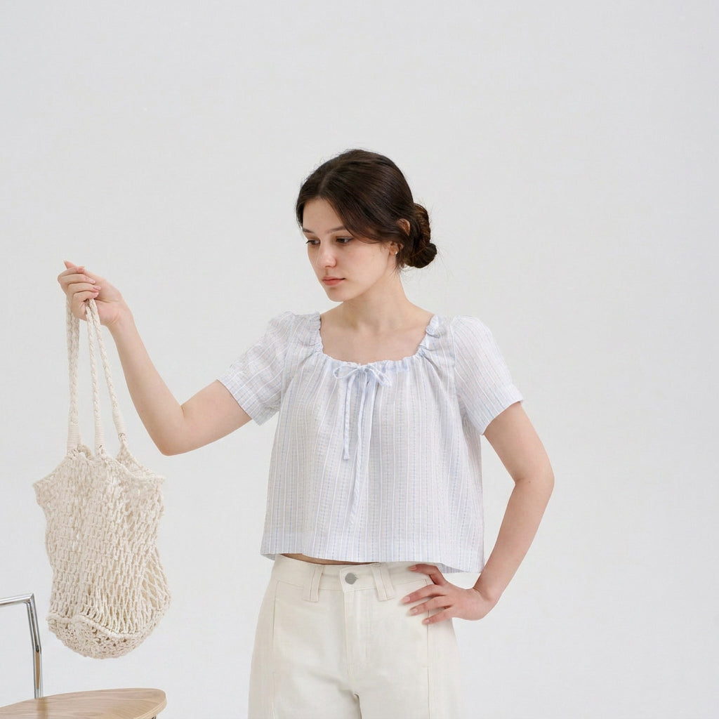 Woman wearing a light blue striped cropped blouse with short sleeves and a front tie detail while holding a cream crochet tote bag.