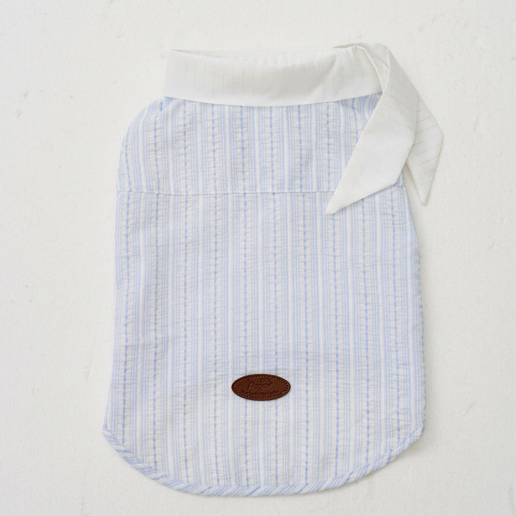 Flat lay of a light blue striped dog shirt with a white collar detail and leather logo patch, designed as small dog apparel and pet clothing for everyday wear.