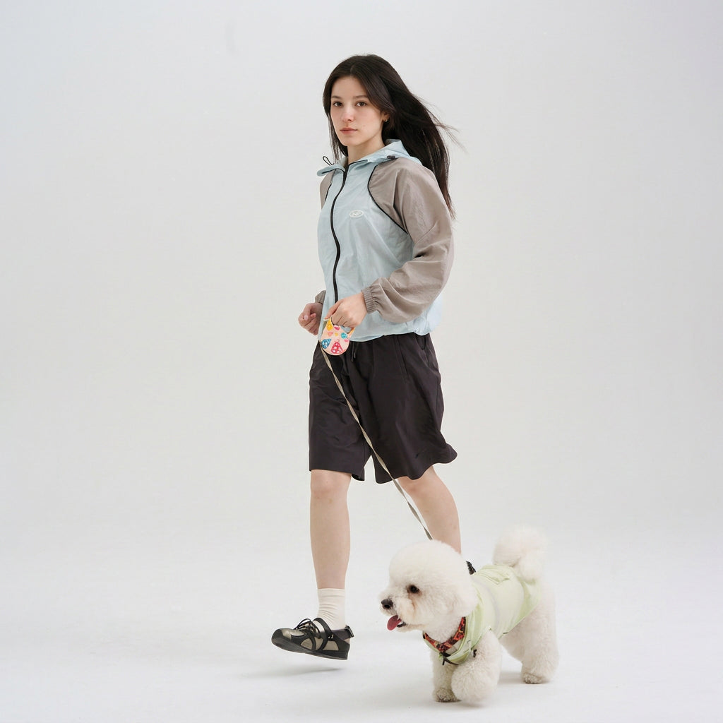 Pet parent wearing a blue and gray sun protection jacket while walking a white small dog in a pale green dog sun jacket, featuring lightweight dog apparel for casual daily walks.