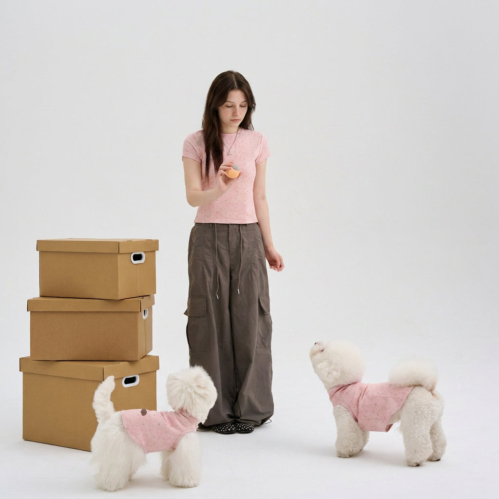 Woman wearing a pink speckled tee standing with two white small dogs in matching pink dog shirts, featuring cute small dog apparel and pet parent matching clothes in a clean studio scene.