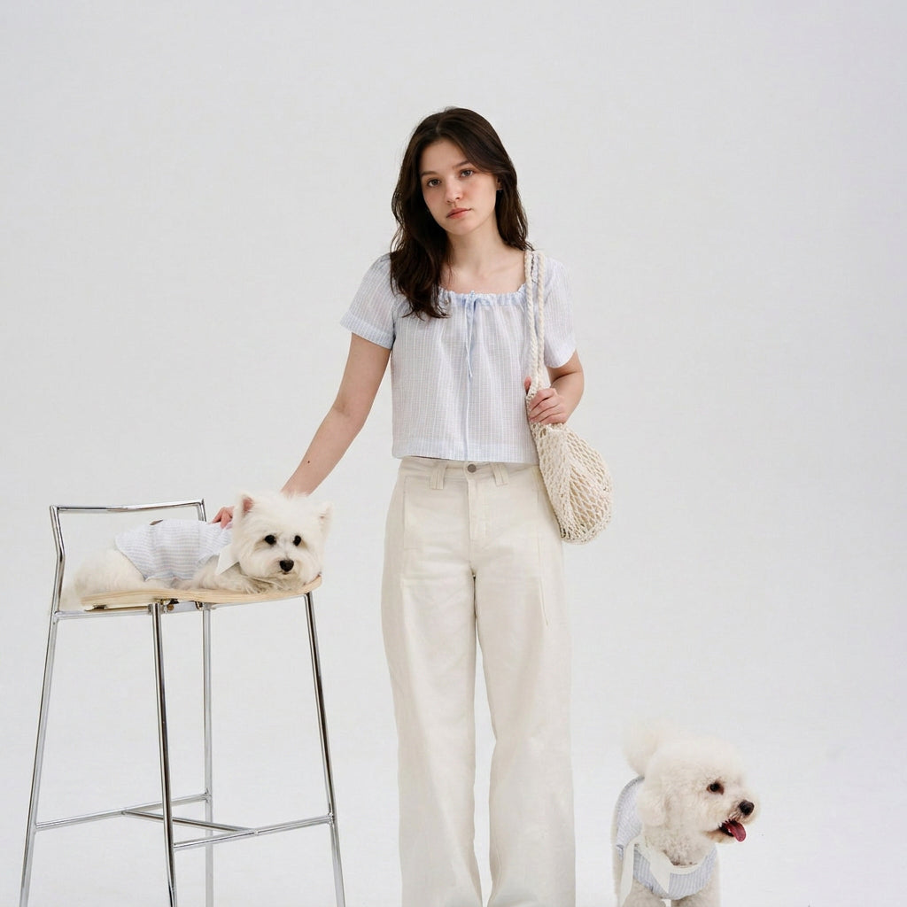 Woman wearing a light blue striped blouse standing with two white small dogs in matching blue striped dog shirts, featuring soft dog apparel and pet parent matching clothes in a clean studio setting.